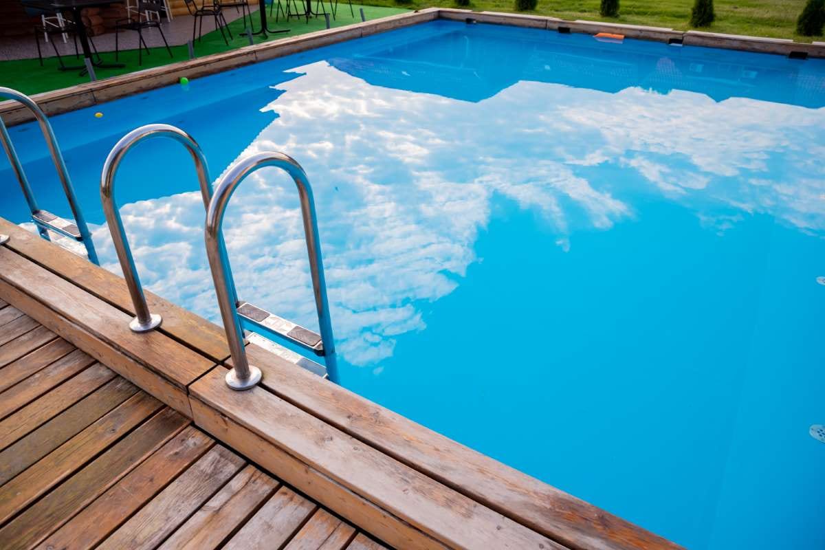Stainless handrails on a pool ladder descending into blue water beside a wooden deck at sunset